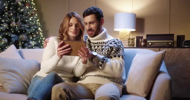 Vidám kaukázusi család pár választott karácsony jelen online tabletta, miközben ül a hangulatos otthon. Férj és feleség karácsonyi ajándékot keresnek a készüléken. Újévi hangulat. Ünnepek — Stock videók