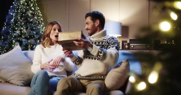 Šťastný překvapený manželka dostává vánoční dárek od milujícího manžela doma na Štědrý den v blízkosti zdobené zářící strom. Běloch dává o svátcích dárek milované ženě. Rodinná oslava — Stock video