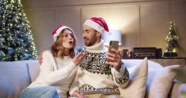 Neşeli aile çifti akıllı telefondan görüntülü sohbet ve el sallarken aynı zamanda parıldayan xmas ağacı beyaz karı ve kocayla evde oturup video konferansında arkadaşlarını selamlıyor.