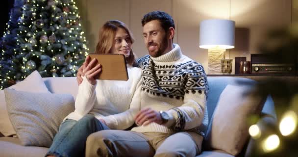 Radostný kavkazský rodinný pár mluví na video chatu on-line na tabletu a mává rukama, zatímco sedí doma s vánoční zdobené strom. Manžel a žena zdraví přátele na videohovoru. Nový rok koncepce — Stock video
