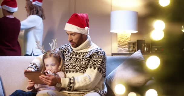 Portrait of happy Caucasian little kid sitting with dad in santa hat at decorated room and tapping on tablet. Mom and teen boy decorating Christmas tree on background. Xmas concept Holiday celebration — Stock Video