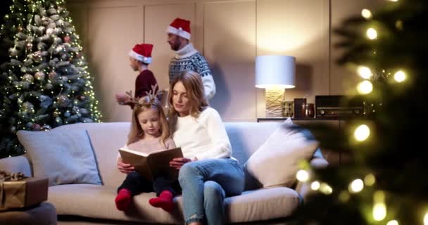美しい白人の幸せなお母さんは、部屋のソファに座って、かわいい娘に読んでいます。背景にクリスマスツリーを飾る父と息子。家族の休日だ。クリスマスの準備だ。明けましておめでとうございます — ストック動画