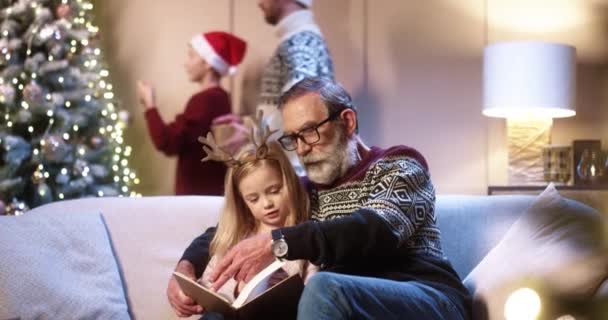 Szczęśliwa, mała, urocza, biała dziewczyna siedząca na kanapie z dziadkiem i czytająca książkę w domu w Wigilię Bożego Narodzenia. Mężczyzna i syn dekorowanie choinki na tle Koncepcja święta Przygotowanie uroczystości — Wideo stockowe
