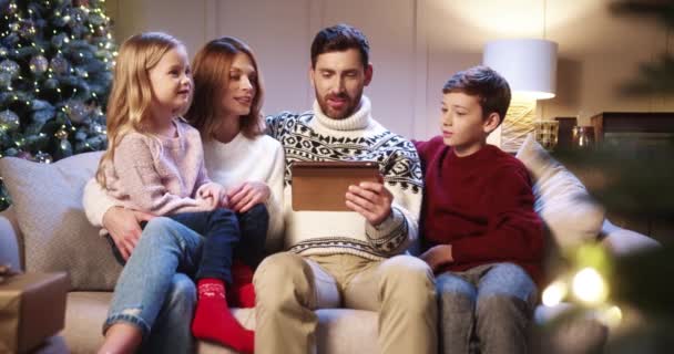 Happy Caucasian family with kids sitting in living cozy room with glowing xmas tree and looking at tablet choosing gifts Parents talking with children on new year eve. Happy holiday concept — Stock Video