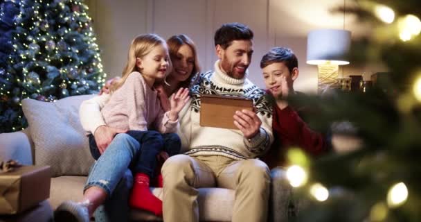 Família caucasiana adorável alegre com crianças sentadas em quarto acolhedor com árvore de xmas brilhante e vídeo conversando em tablet enviando saudações de férias para amigos Pais com crianças falando em videochamada — Vídeo de Stock