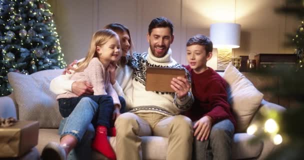 Biała wesoła rodzina z dziećmi siedzącymi w urządzonym pokoju z choinką i wideo na tablecie Rodzice z dziećmi mówiącymi na wideo telefon w Sylwestra. Koncepcja urlopu — Wideo stockowe