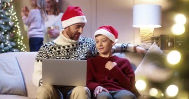 Noel Baba şapkalı mutlu bir beyaz baba ve genç oğlu dizüstü bilgisayarında yazı yazarak Noel ağacının yanında oturuyor. Kış tatilini internetten hediye alarak geçiriyor. Aile tatili konsepti. Yeni Yıl