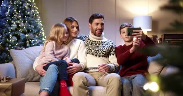 Kaukasische vrolijke familie met kinderen zitten in gezellige ingerichte kamer met kerstboom en het nemen van selfie foto 's op smartphone Zoon het nemen van foto' s met ouders en zus op oudejaarsavond. — Stockvideo