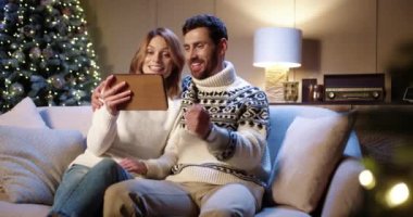 Neşeli gülümseyen beyaz çift, yılbaşı arifesinde Xmas ağacıyla rahat bir odada otururken arkadaşlarıyla tablet üzerinden video konferansında konuşuyor. Karı koca cihazda video konuşuyor.