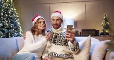 Neşeli gülümseyen beyaz evli çiftin portresi cep telefonuyla sohbet ederken arkadaşlarıyla Noel arifesinde Noel ağacıyla rahat bir odada otururken kutluyor.