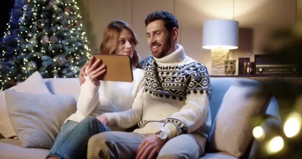Caucasian joyful smiling family couple waving hands and chatting on video call online on tablet with friends while sitting in room with christmas tree on New Years Eve at night Holiday spirit concept — Stock Video