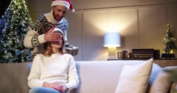 Pareja caucásica enamorada reunida en casa cerca de un árbol de Navidad brillante decorado, el esposo cariñoso hace sorpresa para la esposa cerrando los ojos con las manos dando caja de regalo. Vacaciones de Año Nuevo. De cerca. — Vídeos de Stock