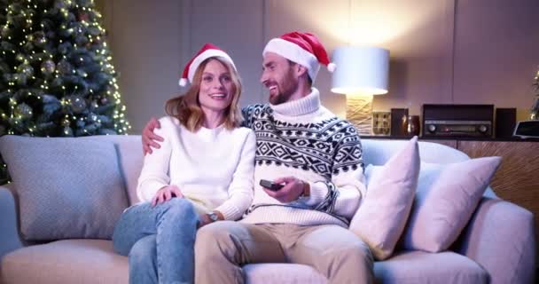 Casamento de família feliz caucasiano casal em chapéus de Papai Noel sentados juntos em casa aconchegante perto de árvore de xmas decorada brilhante e assistindo televisão no Ano Novo. Feliz Natal conceito. Tempo véspera de Natal — Vídeo de Stock