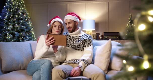 Portret van vrolijke blanke echtpaar man en vrouw rust thuis in de buurt gloeiende nieuwjaarsboom en typen op tablet op zoek naar Kerstmis presenteert online op vakantie verkopen. Gezinsvakantie — Stockvideo