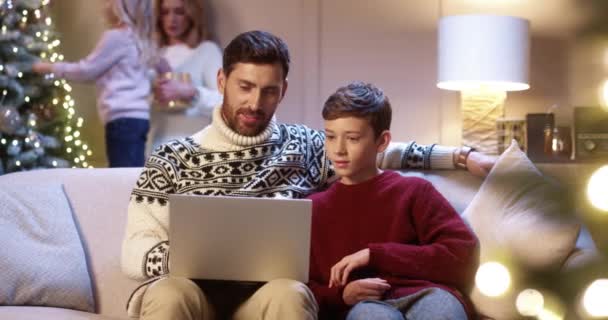 Happy Caucasian vader met tiener zoon zitten in gezellige kamer typen op laptop vakantie tijd doorbrengen op internet kopen geschenken. Mam en klein kind versieren gloeiende kerstboom. Gezinsvakantie — Stockvideo