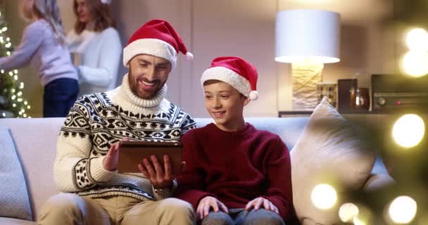 Portrait of joyful Caucasian dad with teen boy sitting in cozy room typing on tablet buying xmas gifts on internet. Mom and small girl decorating glowing Christmas tree. Family holidays concept — Stock Video