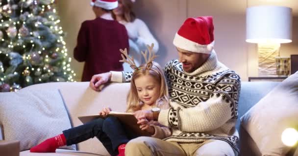 Portrait of joyful caring Caucasian dad with little girl sitting in cozy room at home and reading book. Mom and son decorating glowing Christmas tree. Family holiday time concept. New Year celebration — Stock Video