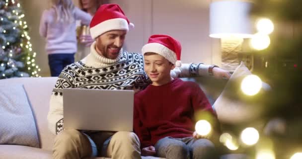 Portret van een liefhebbende blanke vader met een sana hoed en een tienerzoon die thuis zit te browsen op een laptop en vakantietijd doorbrengt met het kijken naar een kerstfilm. Moeder en meisje versieren gloeiende kerstboom vakantie concept — Stockvideo