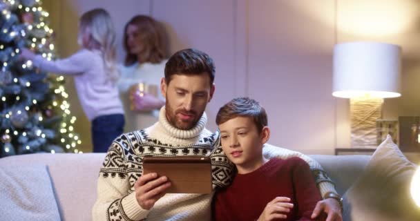 행복 한 백인 아버지와 십 대 아들이 인터넷으로 크리스마스 선물을 고르는 탁자에 앉아 있는 모습, 엄마와 귀여운 딸이 새해 트리를 장식하고 있는 모습 — 비디오