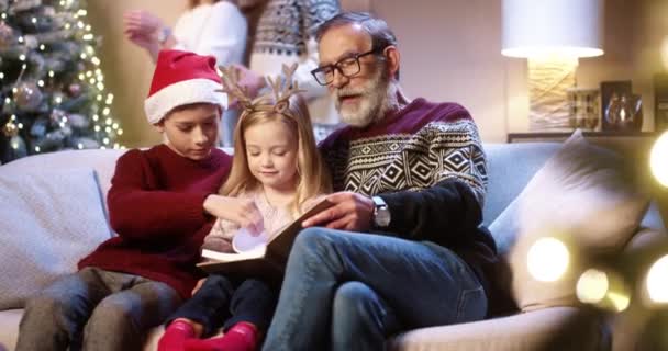 Happy Caucasian family at home on Christmas Eve. Cute kids sitting on sofa with grandpa and reading book while parents decoration xmas glowing tree on background. Happy holidays concept — Stock Video