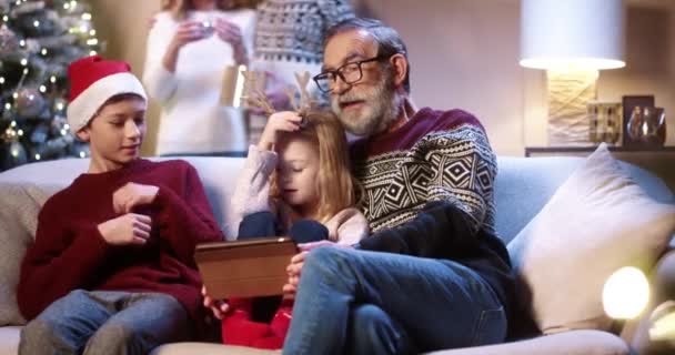 Happy Caucasian family at home on Christmas Eve cute little girl and boy kids with grandpa typing on tablet online while parents decoration xmas glowing tree on background Holidays preparation concept — Stock Video