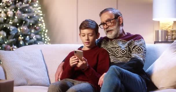 Portret van een gelukkige blanke oude opa met een tienerkleinzoon die in de kamer zit te tikken op de smartphone die kerstcadeau 's kiest op internet in de buurt van gloeiende nieuwjaarsboom. Familie vakantie concept. E-handel — Stockvideo