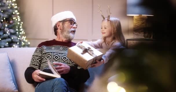 Szczęśliwy biały uroczy dziewczynka i chłopiec dając prezenty dla starego dziadka podczas siedzenia w przytulnym urządzone domu w pobliżu Nowego Roku drzewa i książki do czytania. Dziadek otrzymuje prezenty świąteczne od wnuków — Wideo stockowe
