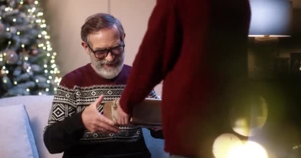 Close up of joyful surprised old grandpa receiving christmas gift from caring teen boy while sitting in decorated room near glowing tree. Grandchild gives xmas present to grandfather. Family concept — Stock Video