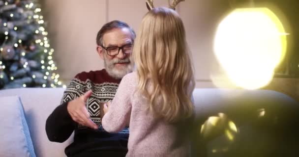 Primer plano retrato de niña linda caucásica feliz dando regalo al viejo abuelo mientras está sentado en casa decorada acogedor cerca de árbol de año nuevo brillante. Abuelo recibir regalo de Navidad de nieto — Vídeo de stock