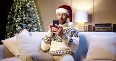 Noel Baba şapkalı neşeli Kafkasyalı yakışıklı adam rahat bir odada Xmas ağacıyla oturuyor ve akıllı telefonda sohbet ediyor tatille tebrik ediyor. - Mutlu yıllar. Kış konsepti Mutlu Noeller.