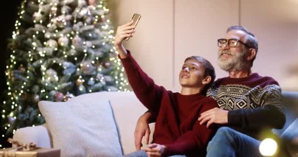 Portret van vrolijke glimlachende blanke oude opa met tienerkleinkind in versierde kamer en het nemen van selfie foto 's op smartphone thuis in de buurt gloeiende nieuwjaarsboom. Kerstvakantie concept — Stockvideo