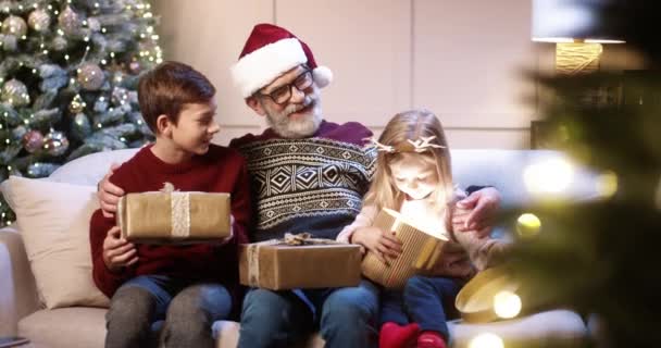 Portrét veselého bělošského rodinného šedovlasého dědečka s vnoučaty otevírajícími vánoční dárky při sezení na pohovce poblíž zdobeného vánočního stromku. Koncept zimních prázdnin. Pocit radosti — Stock video