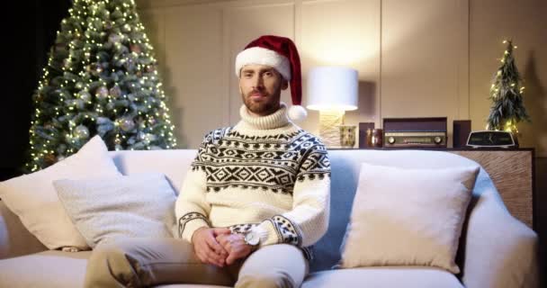 기분좋은 백인 청년이 크리스마스 트리 근처에 있는 장식 된 집에 앉아 있는 기분좋은 모습의 백인 청년은 오직 Xmas Eve 에 불을 반짝이며 앉아 있습니다. 새해 컨셉이야. 12 월 겨울 휴일 — 비디오
