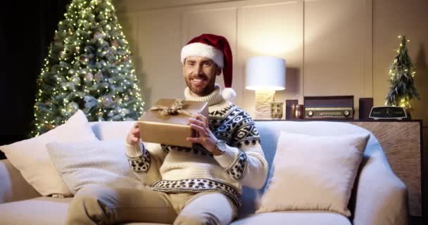 Hombre guapo joven alegre caucásico de buen humor sonriendo con regalo envuelto sentado en la casa decorada cerca del árbol de Navidad con luces centelleantes en Nochebuena. Concepto de Año Nuevo. Vacaciones de diciembre — Vídeos de Stock