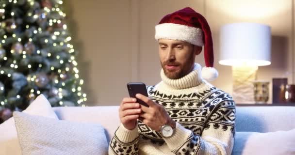 Крупным планом портрет радостного белого красавца в шляпе Санты, сидящего в уютной комнате возле рождественской елки, печатающего на смартфоне с удивленным лицом, выигрывающего рождественскую лотерею. С Новым Годом. Счастливого Рождества! — стоковое видео