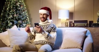 İnternetten xmas alan mutlu, beyaz, yakışıklı bir adam cep telefonuyla ev dekorasyonu yaparak kredi kartıyla ödeme yapıyor. Xmas satışları ve indirimler. Tatil alışverişi. E-ticaret kavramı