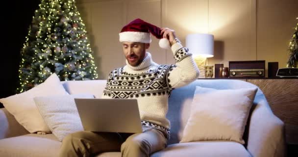 Noel Baba şapkalı mutlu beyaz adamın portresi dekore edilmiş odada oturuyor. Parlayan Xmas ağacının yanında bilgisayardaki görüntülü sohbetten bahsediyor. - Mutlu yıllar. Mutlu Noeller. — Stok video