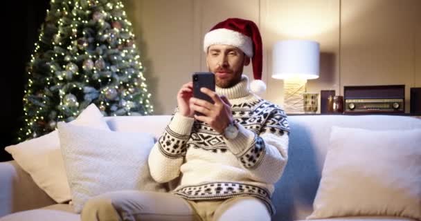 Portret szczęśliwego uśmiechniętego białego młodzieńca w czapce Mikołaja siedzącego na kanapie w urządzonym pokoju, w pobliżu świecącej choinki, mówiącego przez telefon na smartfonie. Szczęśliwego Nowego Roku. Wesołych Świąt — Wideo stockowe