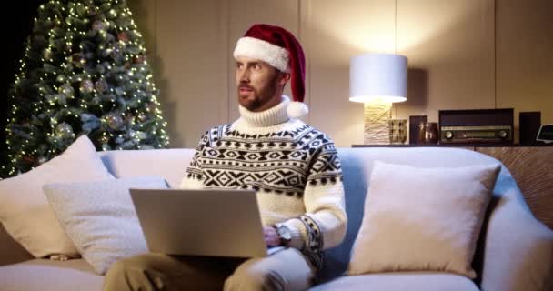 Porträtt av glad vit ung man bär tomte hatt sitter i dekorerade rum nära glödande julgran knacka och bläddra på laptop. Gott nytt år. God jul-konceptet — Stockvideo