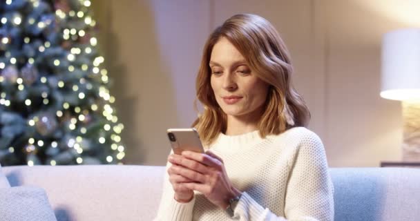 Primer plano retrato de mujer hermosa alegre caucásica con la cara sorprendida sentado en una habitación acogedora cerca de brillante árbol de Navidad escribiendo en el teléfono inteligente. Feliz Año Nuevo. Feliz Navidad. Vacaciones de invierno — Vídeos de Stock