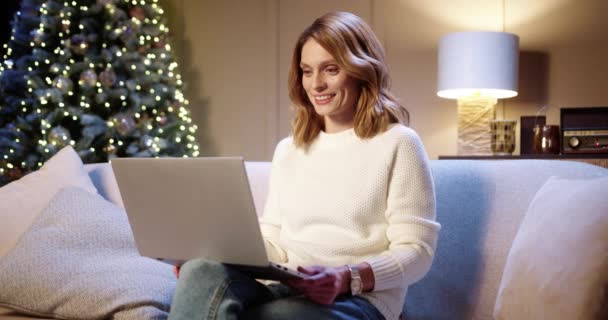밝고 명랑 한 젊은 백인 여성이 크리스마스 트리 근처 소파에 앉아 Xmas Eve 에 있는 노트북으로 비디오 채팅을 하는 모습. 즐거운 휴일 개념 — 비디오