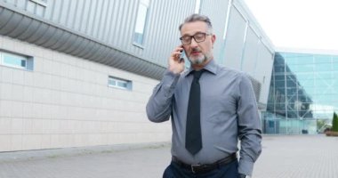 Başarılı, gözlüklü ve kravatlı beyaz iş adamı dışarıda yürüyor ve cep telefonuyla konuşuyor. İş merkezinin yanındaki sokakta cep telefonuyla konuşan yakışıklı bir kıdemli. Telefon görüşmesi.