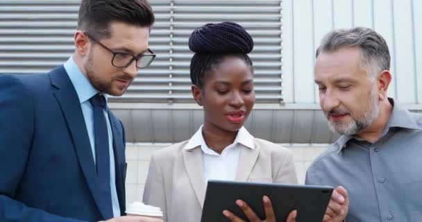 Jovem empresária afro-americana e dois empresários caucasianos discutindo o trabalho com tablet nas mãos na rua. Mixed-corridas mulher e homens falando ao ar livre Olhando para tela gadget. — Vídeo de Stock