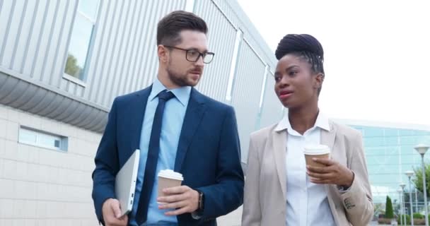 Razze miste giovane uomo d'affari e donna d'affari a piedi all'aperto, tenendo caffè-to-go e parlando. Uomo d'affari multietnico e donna d'affari passeggiando per strada e discutendo di affari con bevande. — Video Stock