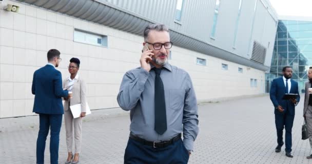 戴眼镜打着领带站在户外用手机聊天的白人商人。一位英俊的老人在商业中心附近的街上用手机说话。混血儿. — 图库视频影像