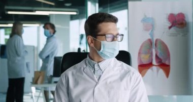 Tıbbi maske takan yakışıklı beyaz erkek doktorun, iş yerindeki klinikte kameraya bakıp gülümseyen portresi. Koronavirüs salgını. Hastane kavramı