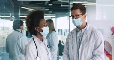 Kafkas erkek meslektaşı ile birlikte duran ve Coronavirus salgını sırasında klinikteki kameraya bakan mutlu Afrika kökenli Amerikalı kadın doktor portresi. Sağlık kavramı