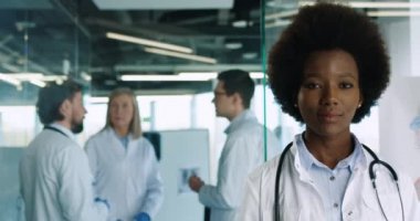 Hastanede kameraya gülümseyen genç Afro-Amerikan profesyonel doktor portresini yakından çek. Beyaz doktorlar arka planda konuşuyor. Sağlık hizmetleri kavramı