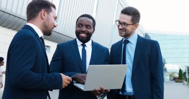Empresários afro-americanos e caucasianos em pé na rua, sorrindo e discutindo o trabalho com computador portátil nas mãos. Homens de raças mistas parceiros falando sobre projeto de trabalho. — Vídeo de Stock