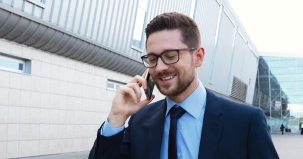 Kavkazský pohledný mladý obchodník v obleku, kravatě a brýlích, jak mluví na smartphonu a venku. Hezký muž v obchodním stylu mluví na mobilním telefonu. Rozhovor.